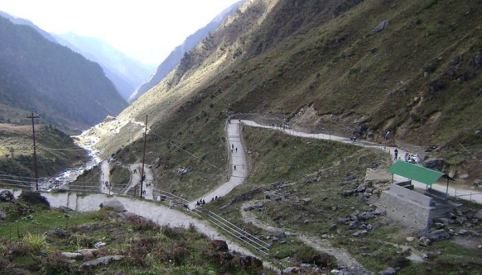 A winding mountain road leading towards Kedarnath Temple, surrounded by lush greenery and snow-capped peaks