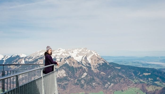 Joyful tourist woman standing with hands up in achievement at Stanserhorn Summit, Engelberg, Switzerland, with snow-covered mountains in the background